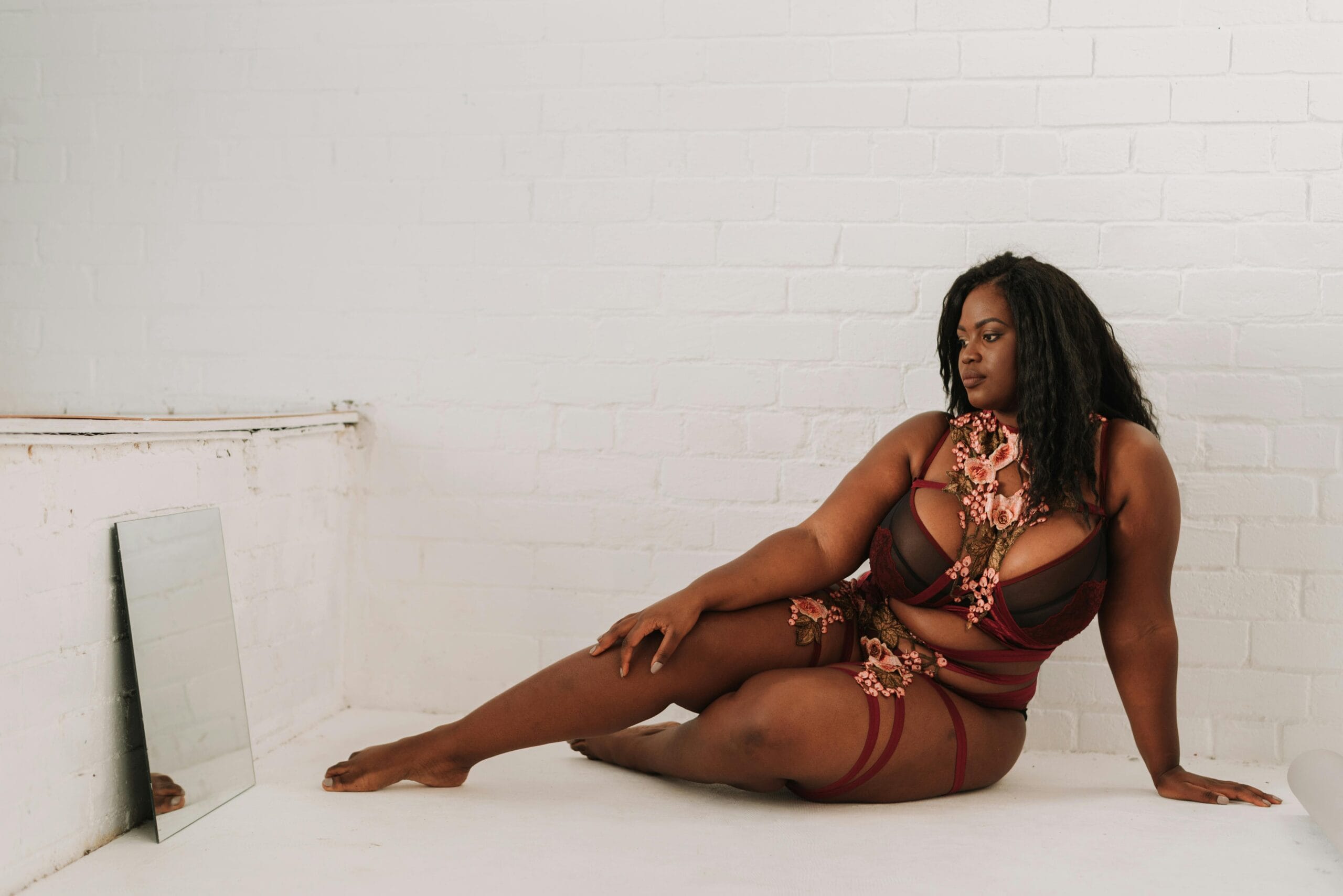 Full body of gorgeous barefoot overweight African American female in underwear leaning on floor and looking at reflection in mirror